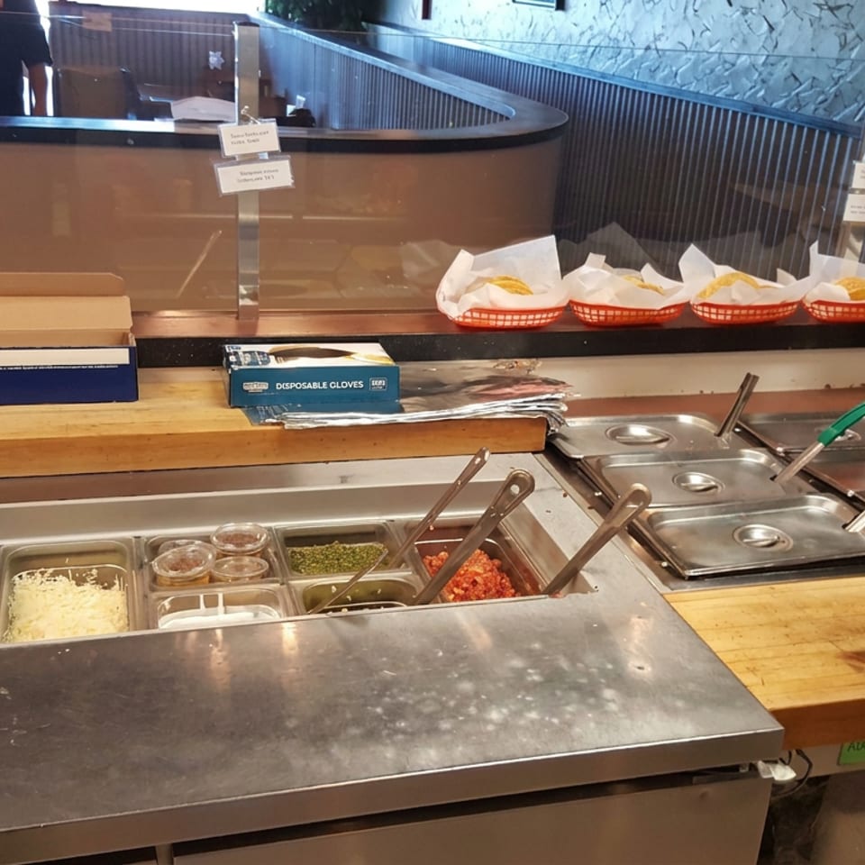 Restaurant self-service counter with baskets of prepared food, ingredient trays, serving utensils, gloves, and a condiment station.