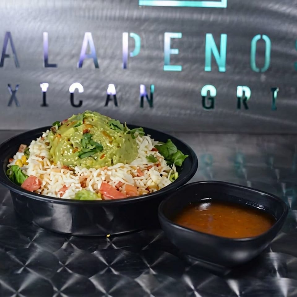 Mexican grill bowl topped with guacamole, rice, lettuce, tomatoes, and crispy tortilla strips beside a bowl of orange sauce.