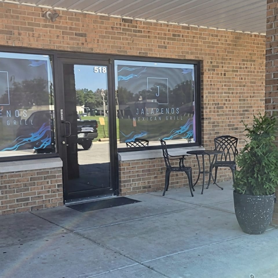 Exterior entrance of Jalapenos Mexican Grill with branded windows, brick walls, patio table, chairs, and potted greenery.