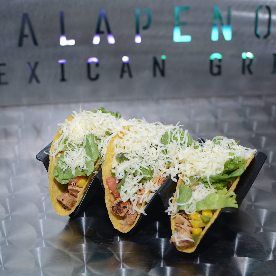 Three hard-shell tacos filled with grilled meat, lettuce, corn, tomatoes, and shredded cheese on a black tray at a Mexican grill.
