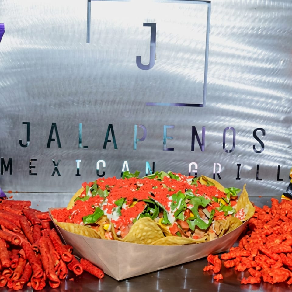 Loaded nachos topped with red seasoning, cheese, and cilantro, surrounded by Takis Fuego and Flamin’ Hot Cheetos bags and snacks.