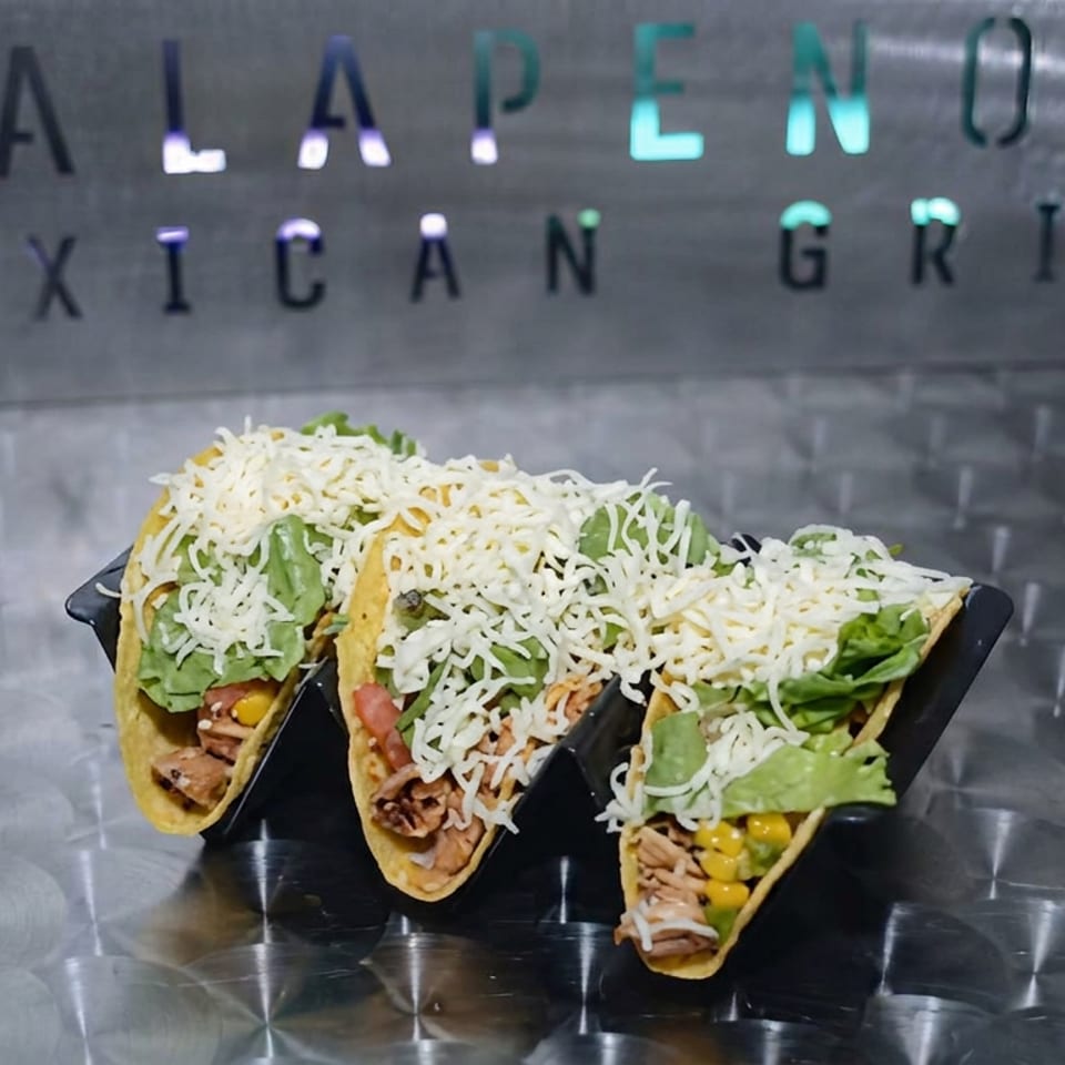 Three hard-shell tacos filled with meat, lettuce, corn, and shredded cheese sit in a black holder on a metallic counter.