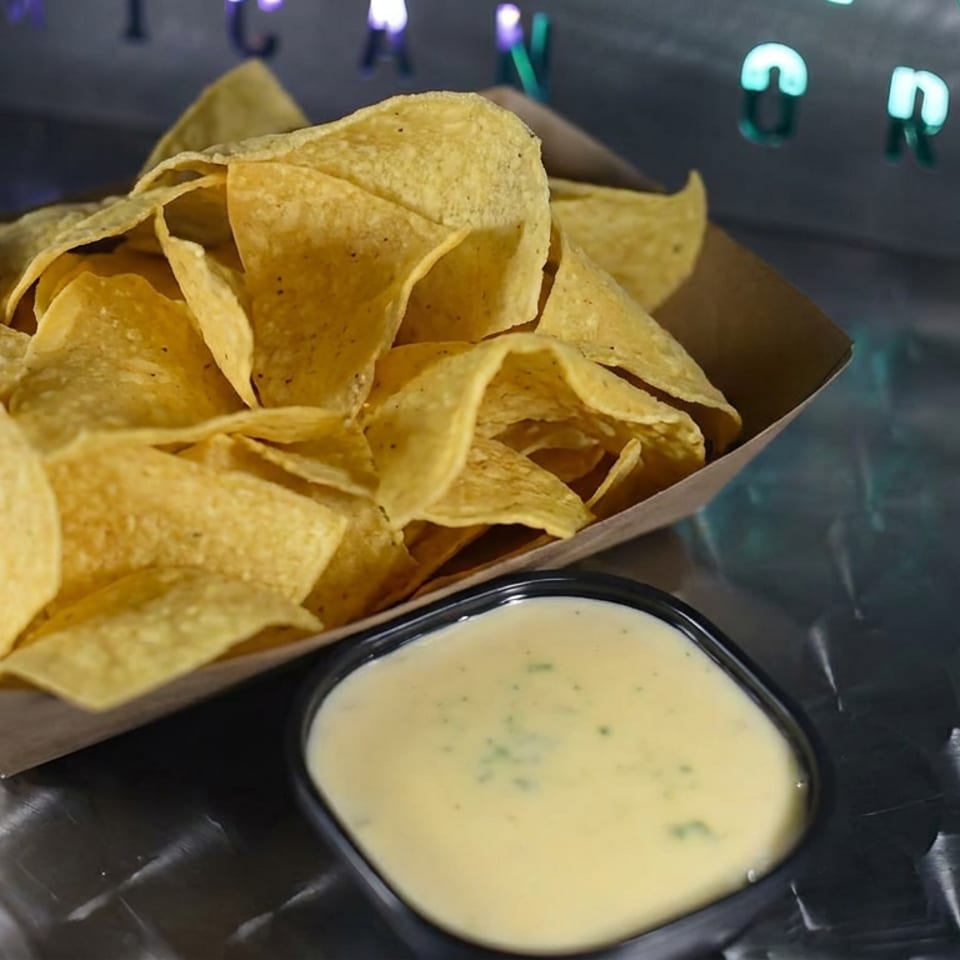 Crispy tortilla chips in a paper tray served with creamy cheese dipping sauce on a reflective restaurant table.