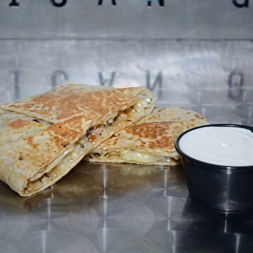 Grilled cheese quesadillas stacked beside a cup of creamy white dipping sauce on a reflective metal surface.