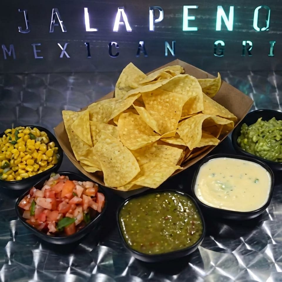 Tortilla chips surrounded by bowls of corn salsa, pico de gallo, green salsa, queso, and guacamole beneath a Jalapeño Mexican Grill sign.