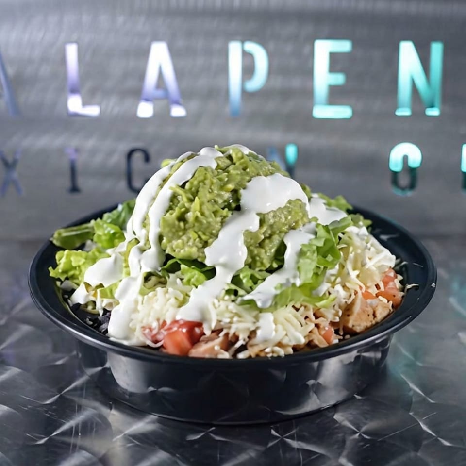 Black bowl of chopped salad topped with guacamole, shredded cheese, tomatoes, lettuce, and creamy white dressing on a metal counter.